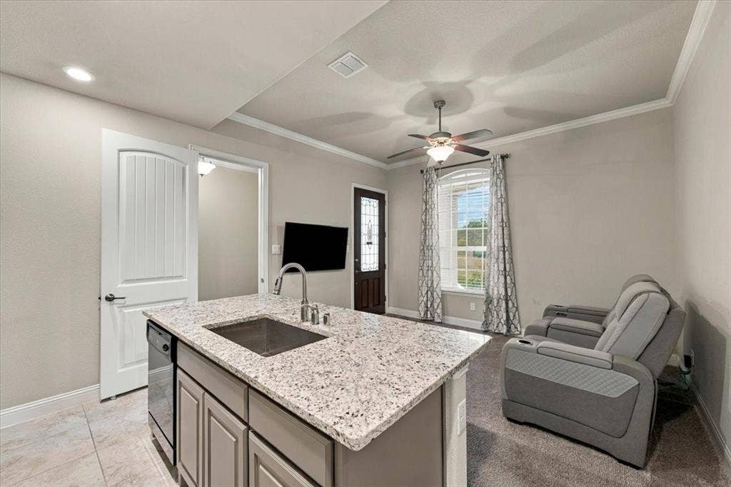 1455 Corrara Drive, Rockwall, TX 75032 Suite Kitchenette features an island with an undermount sink, built-in dishwasher, and cabinet storage, with an open layout to the sitting room. Ceramic tile flooring extends throughout, transitioning to carpet in the sitting room.