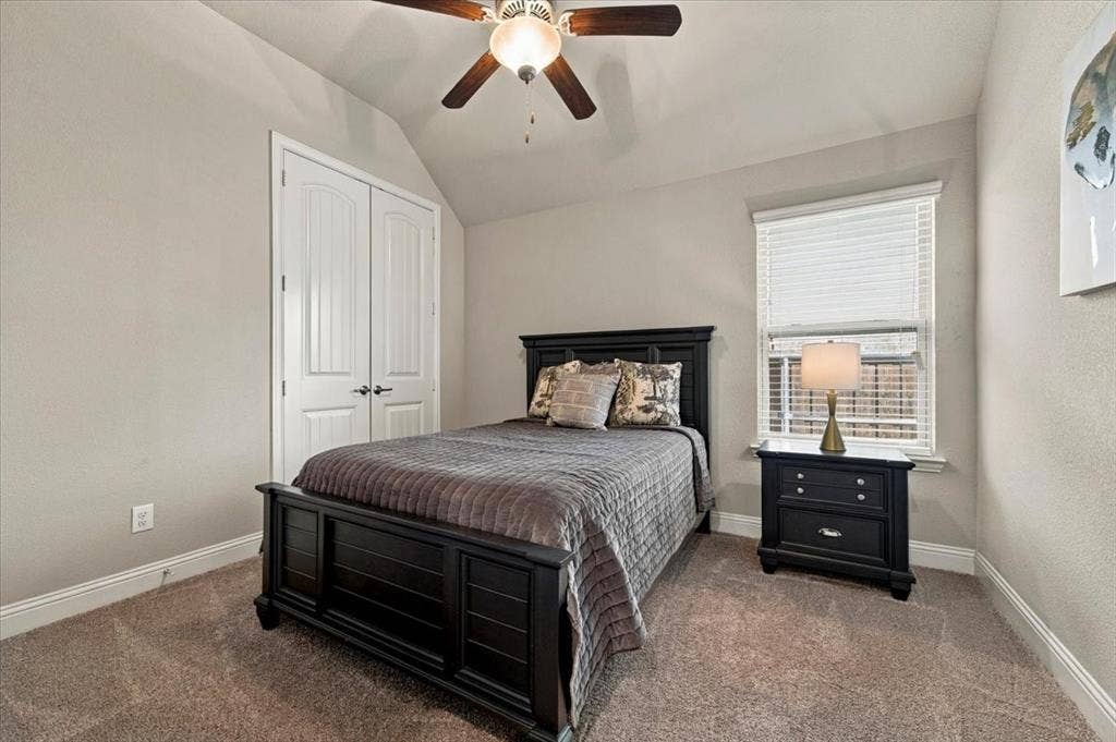 1455 Corrara Drive, Rockwall, TX 75032 Secondary Bedroom features a vaulted ceiling and a ceiling fan with light fixture, a window, and a double-door closet. Carpet flooring extends throughout.