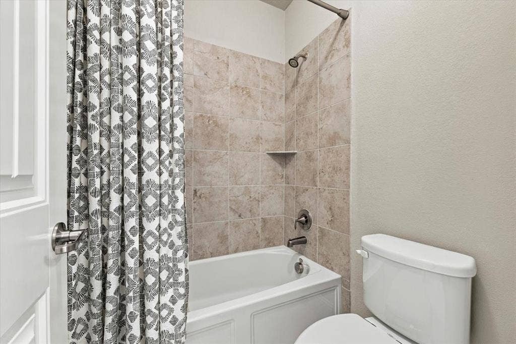 1455 Corrara Drive, Rockwall, TX 75032 Secondary Bathroom features a separate compartment with a tub/shower combination, tile surround with a corner shelf, and a toilet. Ceramic tile flooring extends throughout.