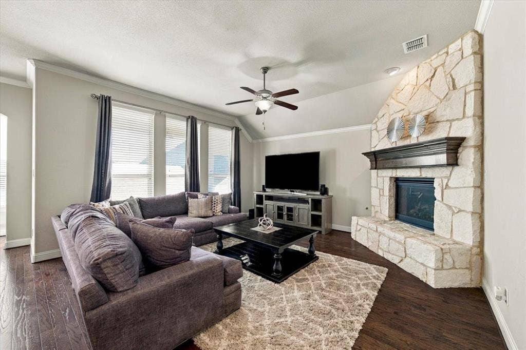1455 Corrara Drive, Rockwall, TX 75032 Living Room features an angled ceiling section, a gas log fireplace with gas starter, floor-to-ceiling stonework and a raised stone hearth, and a row of three windows. Wood flooring extends throughout.
