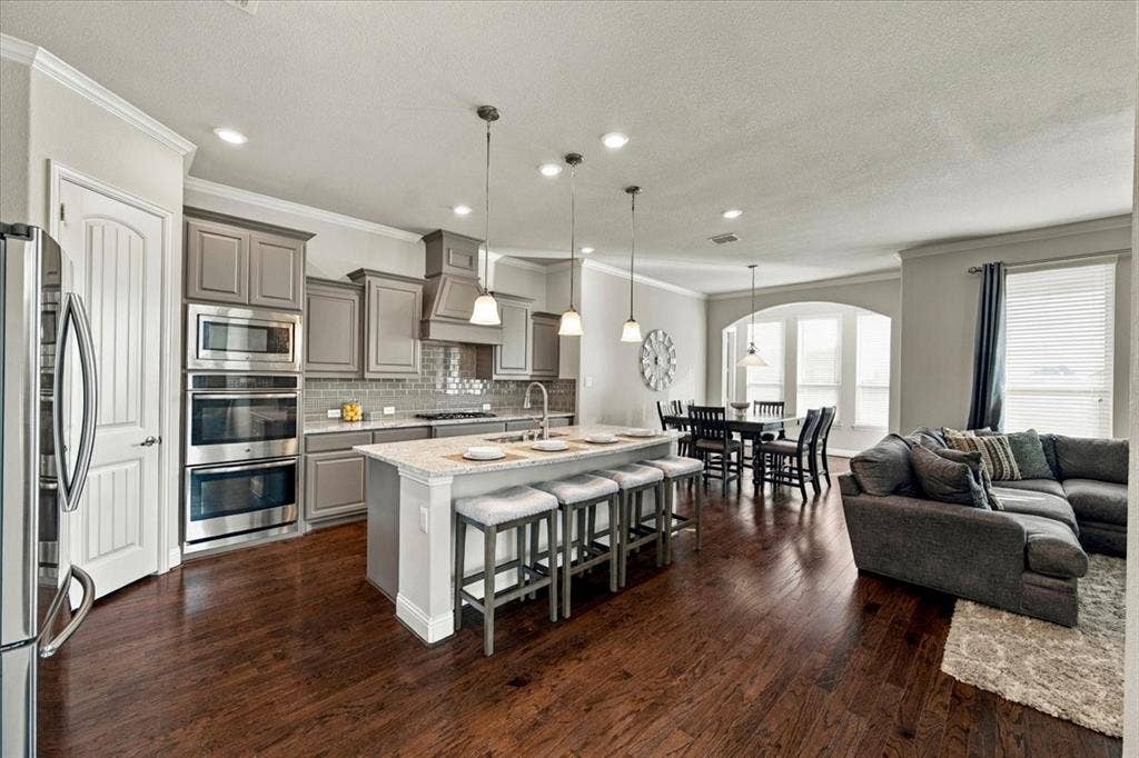 1455 Corrara Drive, Rockwall, TX 75032 Kitchen features recessed lighting that continues into the dining area, island seating, and an open layout connecting the kitchen, dining area, and living room. Wood flooring extends throughout.