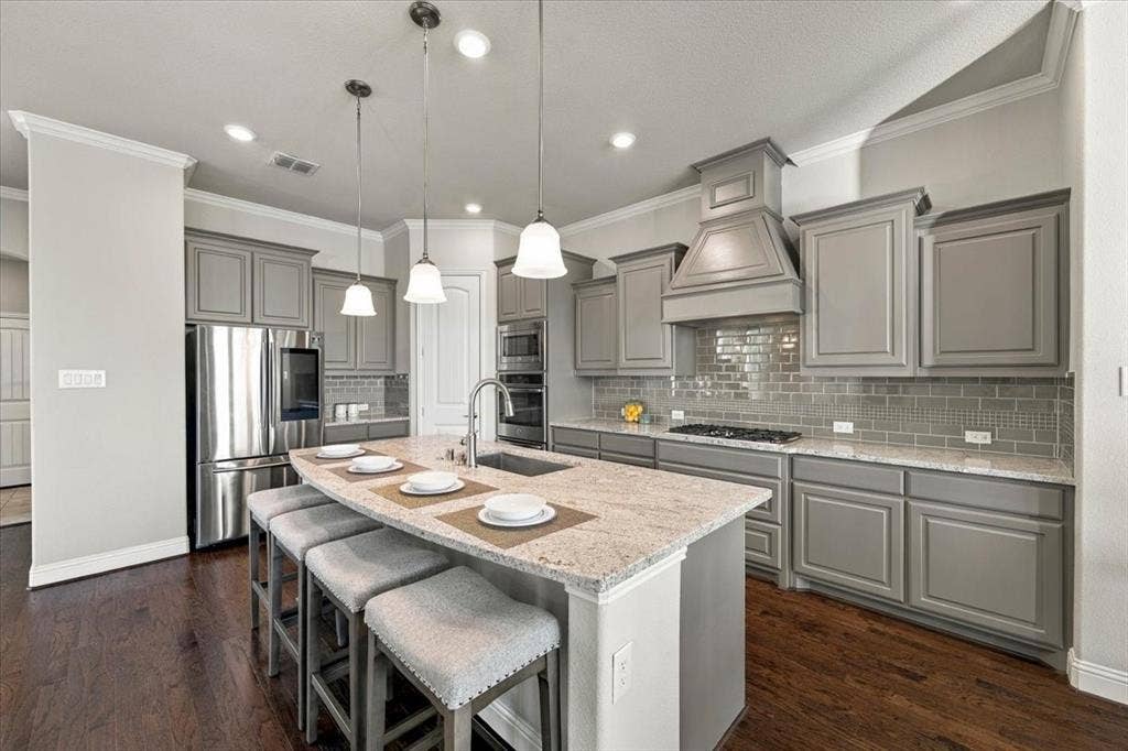 1455 Corrara Drive, Rockwall, TX 75032 Kitchen features crown molding, three pendant lights over the island, a decorative vent hood, upper cabinets with crown trim, and a curved island with breakfast bar seating. Wood flooring extends throughout.