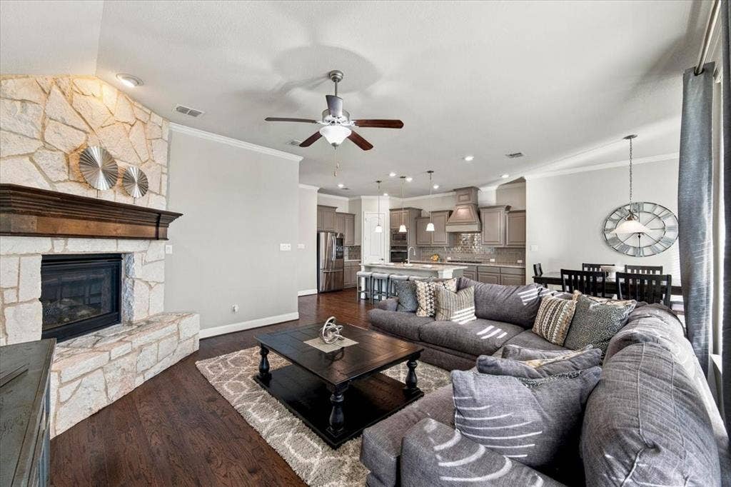 1455 Corrara Drive, Rockwall, TX 75032 Living Room features crown molding, a ceiling fan with light fixture, a corner gas log fireplace with gas starter, a stone surround and wood mantel, and an open layout connecting to the kitchen and dining area. Wood flooring extends throughout.