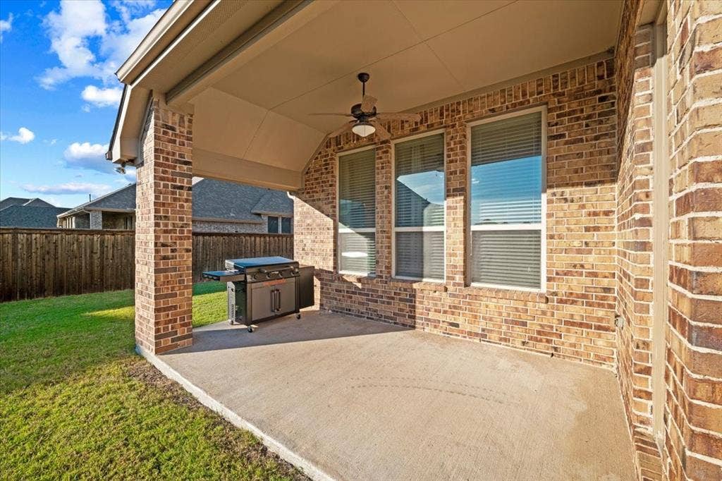 1455 Corrara Drive, Rockwall, TX 75032 Covered Patio features a ceiling fan with light fixture, brick columns and walls, three windows, and a view of the fenced backyard. Concrete flooring extends throughout.