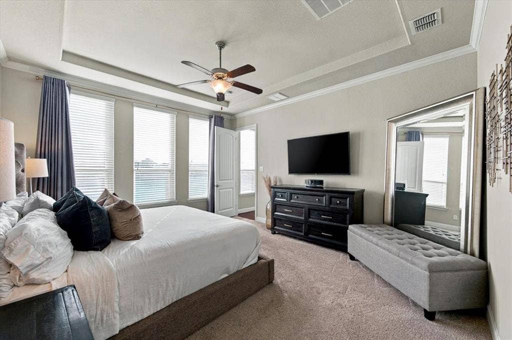 1455 Corrara Drive, Rockwall, TX 75032 Primary Bedroom features a tray ceiling with crown molding, a ceiling fan with light fixture, a row of three windows, and an entry door. Carpet flooring extends throughout.