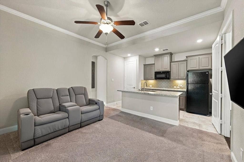 1455 Corrara Drive, Rockwall, TX 75032 Suite Sitting Room features crown molding and a ceiling fan with light fixture, an arched opening to the suite hall, and an open layout to the kitchenette. Carpet flooring extends throughout, transitioning to ceramic tile in the kitchenette.