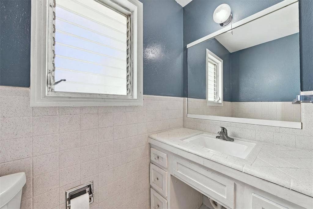 White vanity featuring an integrated sink, tile countertop, large wall-mounted mirror, louvered window, and chrome finish faucet