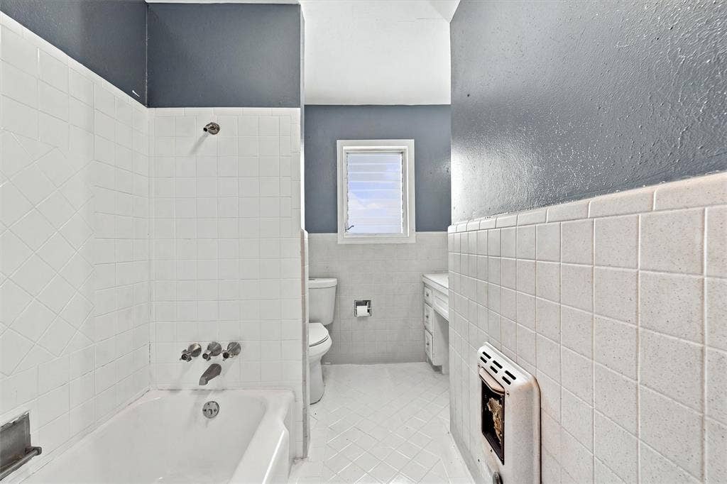 Bathroom featuring a white porcelain tub with integrated shower fixture, white ceramic wall tile, white tiled flooring, and a window with louvered glass panes