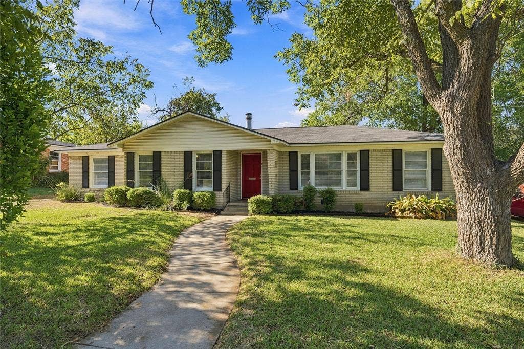 Single-story brick exterior featuring a prominent red front door, contrasting shutters, a paved walkway, and mature landscape elements