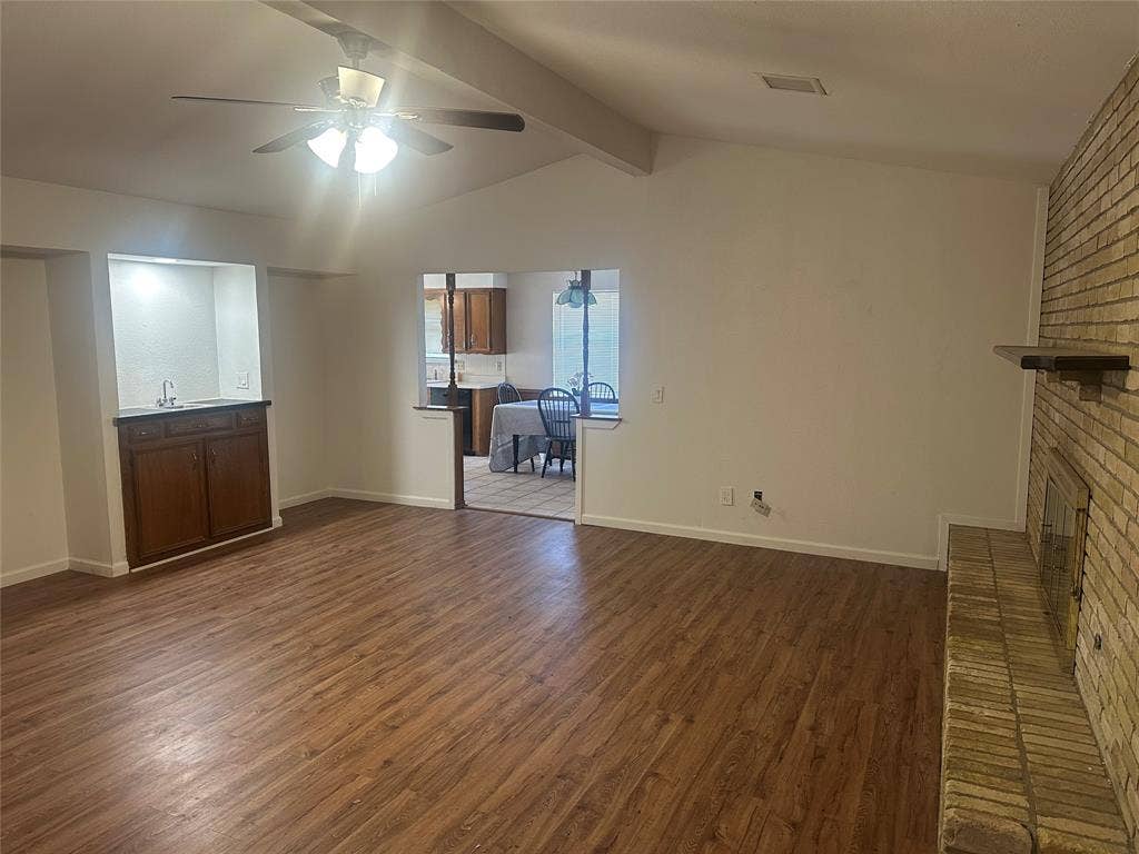 Vaulted ceiling with exposed beam, wood-finish flooring, full-height masonry fireplace, and built-in wet bar featuring dark wood cabinetry and a sink