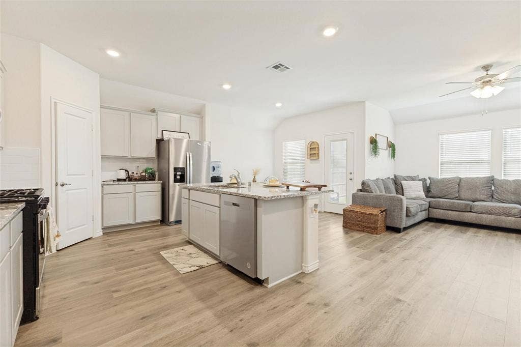 Open-concept kitchen featuring shaker-style cabinetry, stone countertops, a central island with an undermount sink, stainless steel appliances, and wood-finish flooring throughout