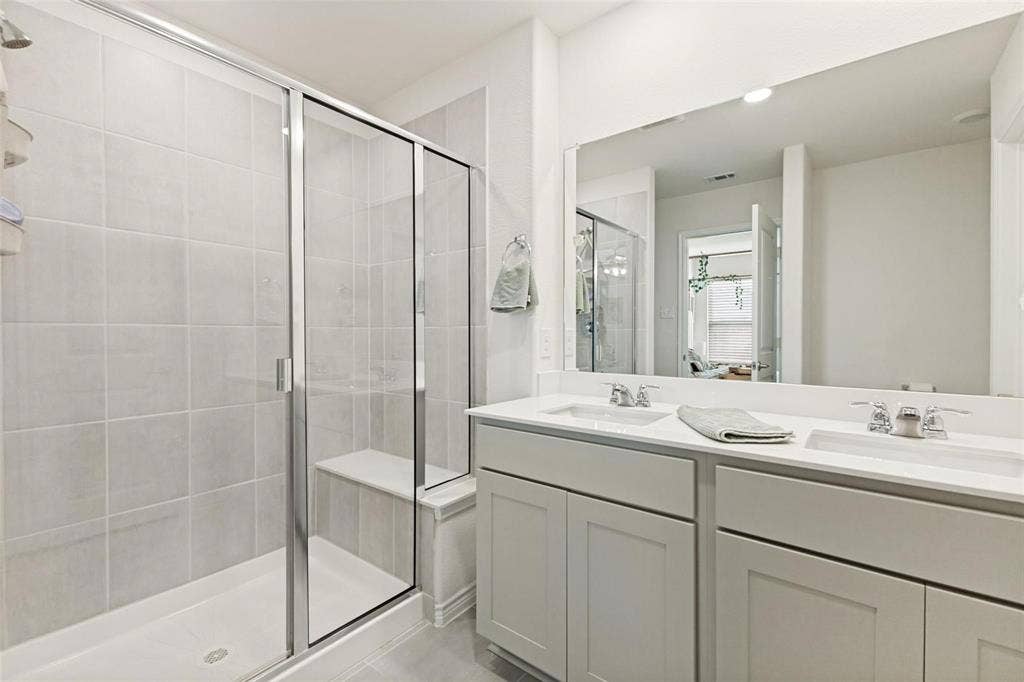 Bathroom featuring a dual-sink vanity with solid-surface countertop, full-width mirror, and a walk-in shower with tiled surround and integrated bench