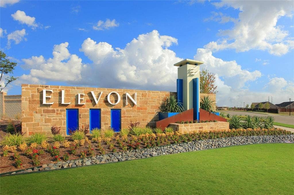 Stone-veneer community monument featuring structural accent pillars, integrated lighting, metal detailing, and tiered ornamental landscaping