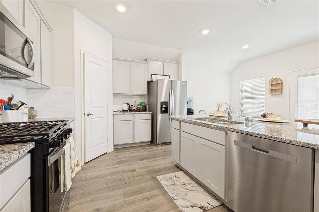 Kitchen featuring light-toned shaker cabinetry, stone-finish countertops, stainless steel appliances, a center island with an integrated double basin sink, and wood-finish flooring