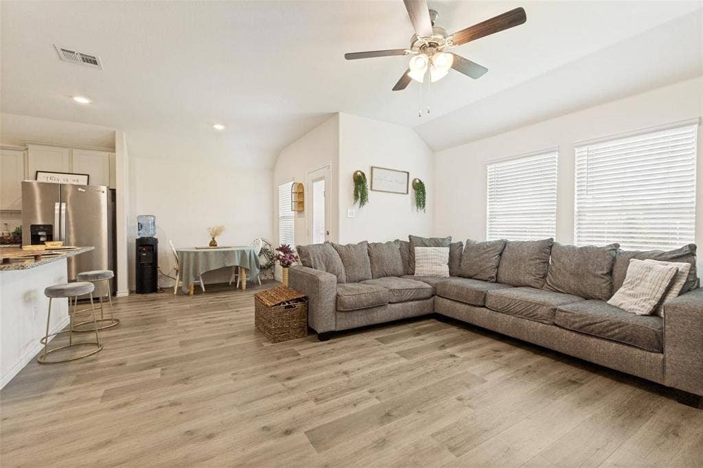 Open-concept interior featuring wood-finish flooring, a vaulted ceiling with a central paddle fan, stone-surface breakfast bar, and stainless steel refrigerator