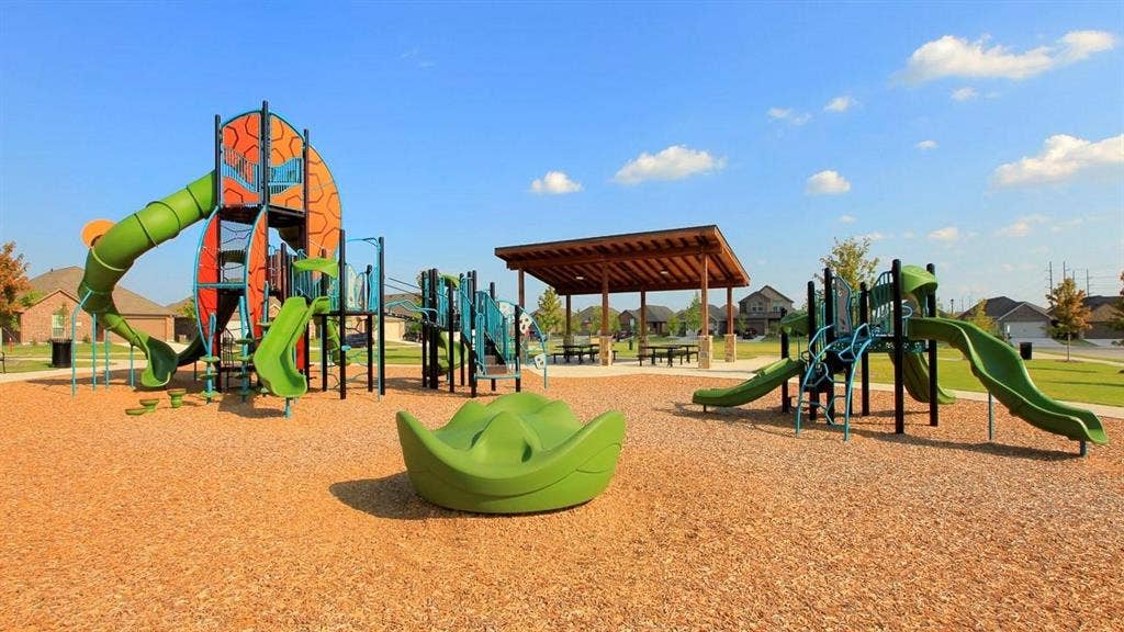 Community playground featuring multiple modular climbing structures, green slides, a central rotating element, and a covered timber-framed pavilion