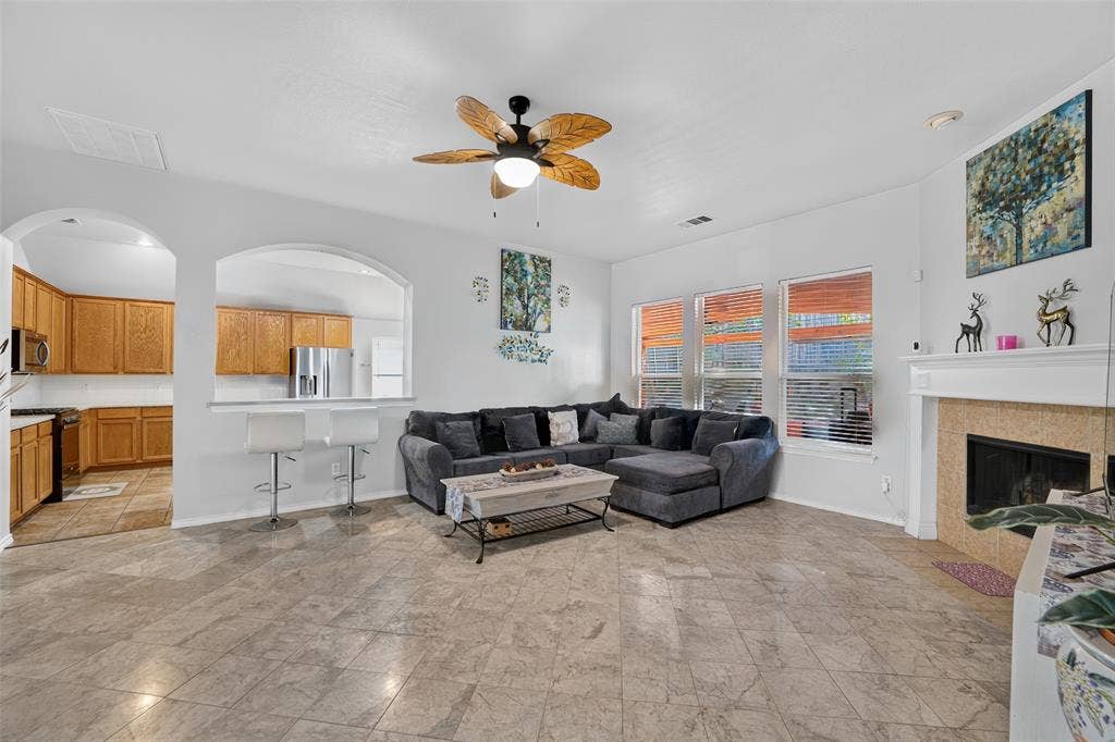 Open-concept living space featuring stone-finish tile flooring, a fireplace with a traditional mantel, an arched pass-through window, and a ceiling fan with leaf-style blades