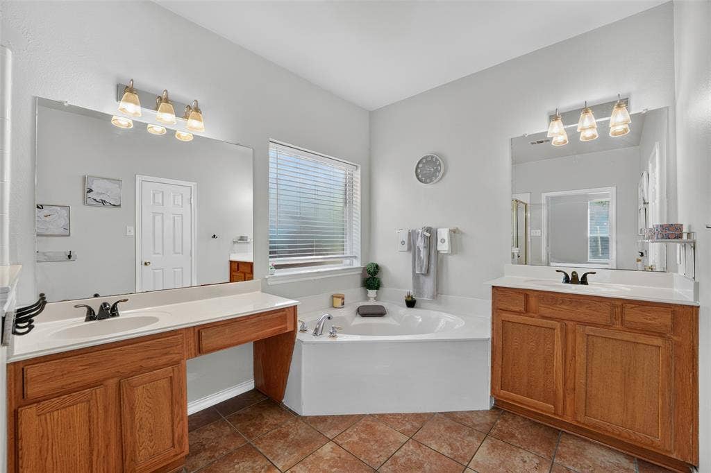 Dual wood-finish vanities with light-toned countertops, corner soaking tub, ceramic tile flooring, and large wall-mounted mirrors