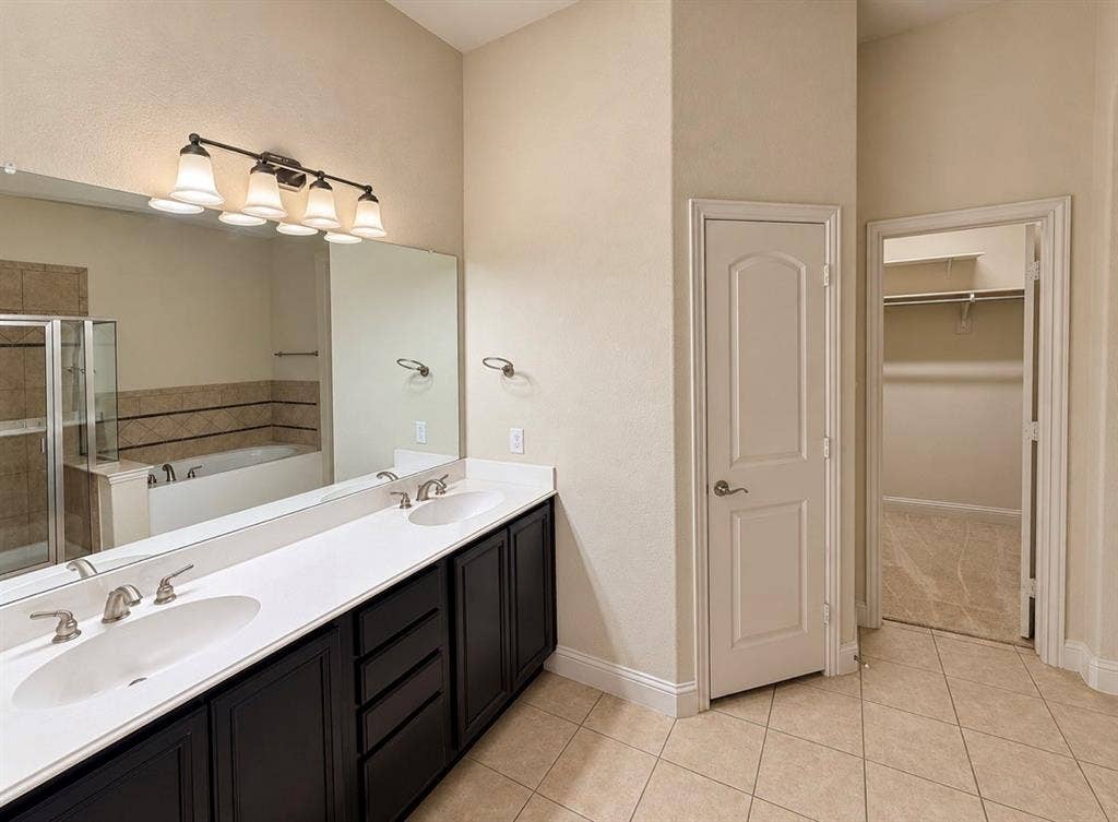 8616 Naomi Street, Plano, TX 75024 Dual-sink vanity with white countertops and dark-finish cabinetry, full-width wall mirror, four-light vanity fixture, built-in soaking tub, and tile flooring