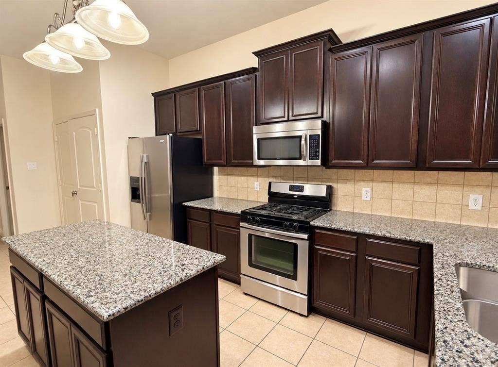 8616 Naomi Street, Plano, TX 75024 Kitchen featuring dark wood cabinetry, speckled stone-finish countertops, stainless steel appliances, a central island, and tile flooring
