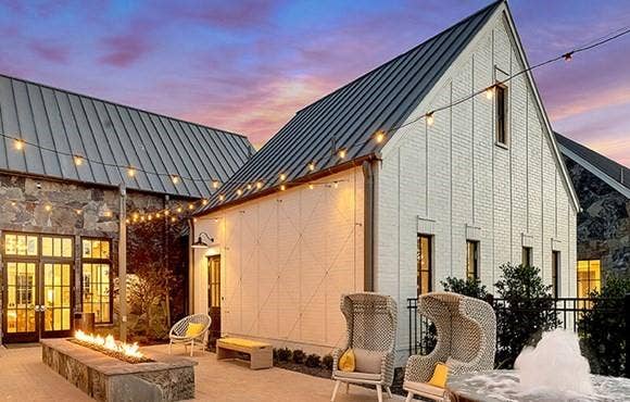 9096 Phoebe Road, Frisco, TX 75035 Modern farmhouse exterior featuring vertical white board-and-batten siding, stone masonry accents, and a dark metal roof, complemented by a paver patio with a built-in linear stone fire feature