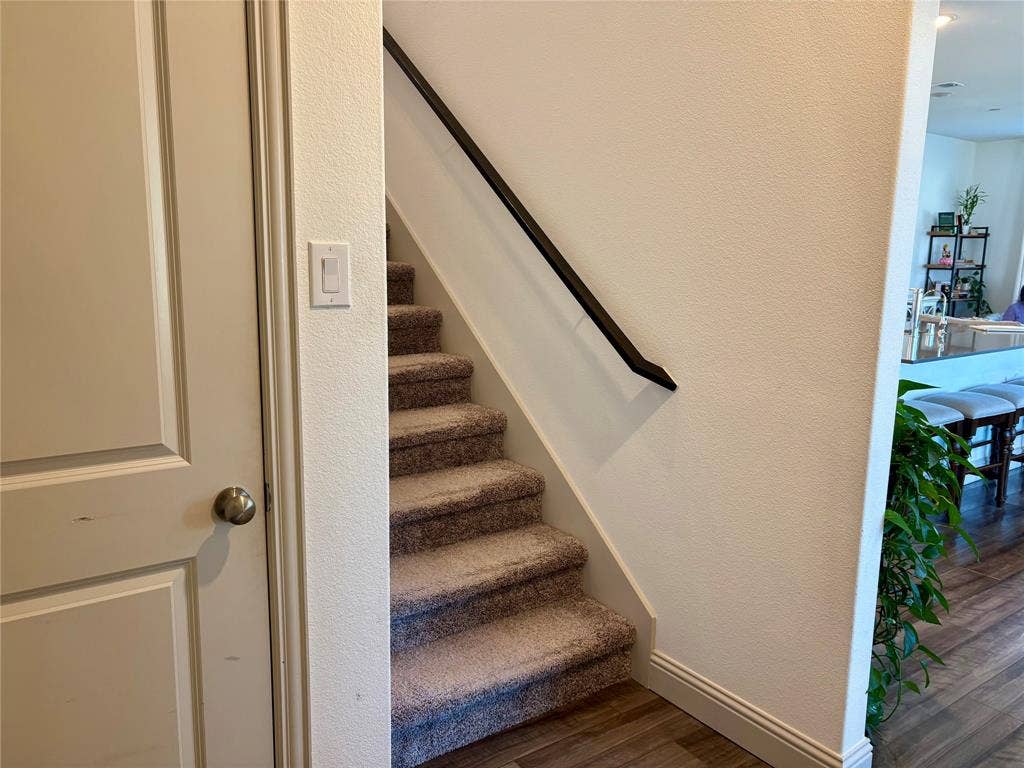 9096 Phoebe Road, Frisco, TX 75035 Staircase with neutral-toned carpeting and a dark-finished handrail, accompanied by a solid-panel door and wood-finish flooring