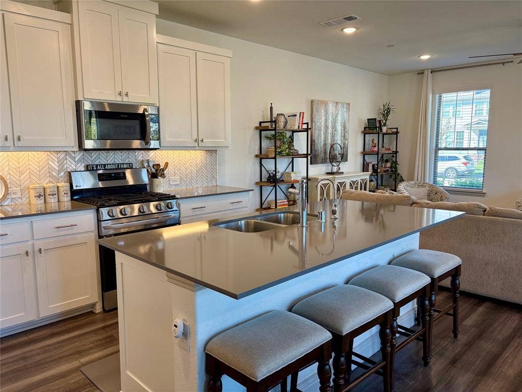 9096 Phoebe Road, Frisco, TX 75035 Kitchen featuring a large central island with a solid surface countertop, white shaker-style cabinetry, a herringbone-pattern tile backsplash, stainless steel appliances, and wood-finish flooring