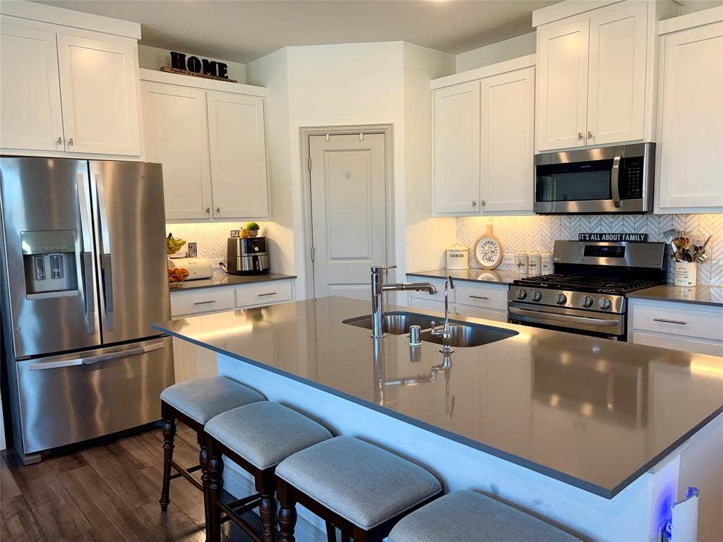 9096 Phoebe Road, Frisco, TX 75035 Kitchen featuring white shaker-style cabinetry, stainless steel appliances, a central island with a solid-surface countertop, and a herringbone-patterned tile backsplash