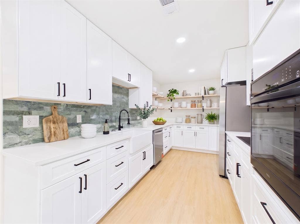 Kitchen featuring white cabinetry with matte black hardware, solid surface countertops, a farmhouse-style apron sink, a glass tile backsplash, and built-in stainless steel appliances