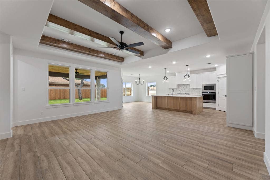 Unfurnished living room featuring baseboards, beamed ceiling, light wood-style floors, and ceiling fan with notable chandelier