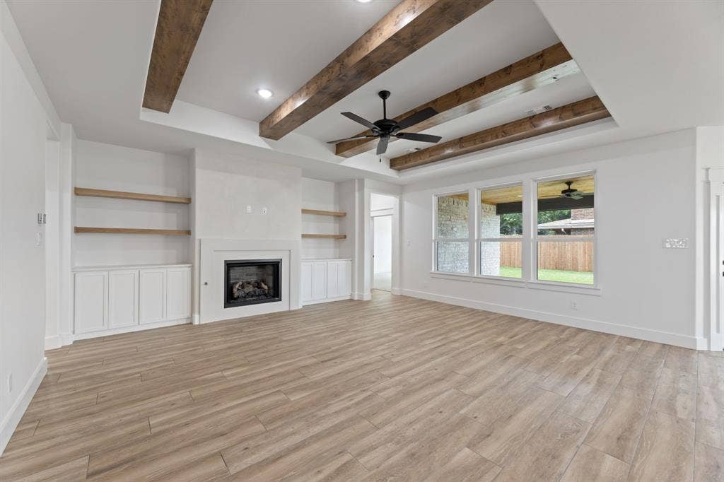 Unfurnished living room featuring light wood-style flooring, baseboards, ceiling fan, a fireplace, and beam ceiling