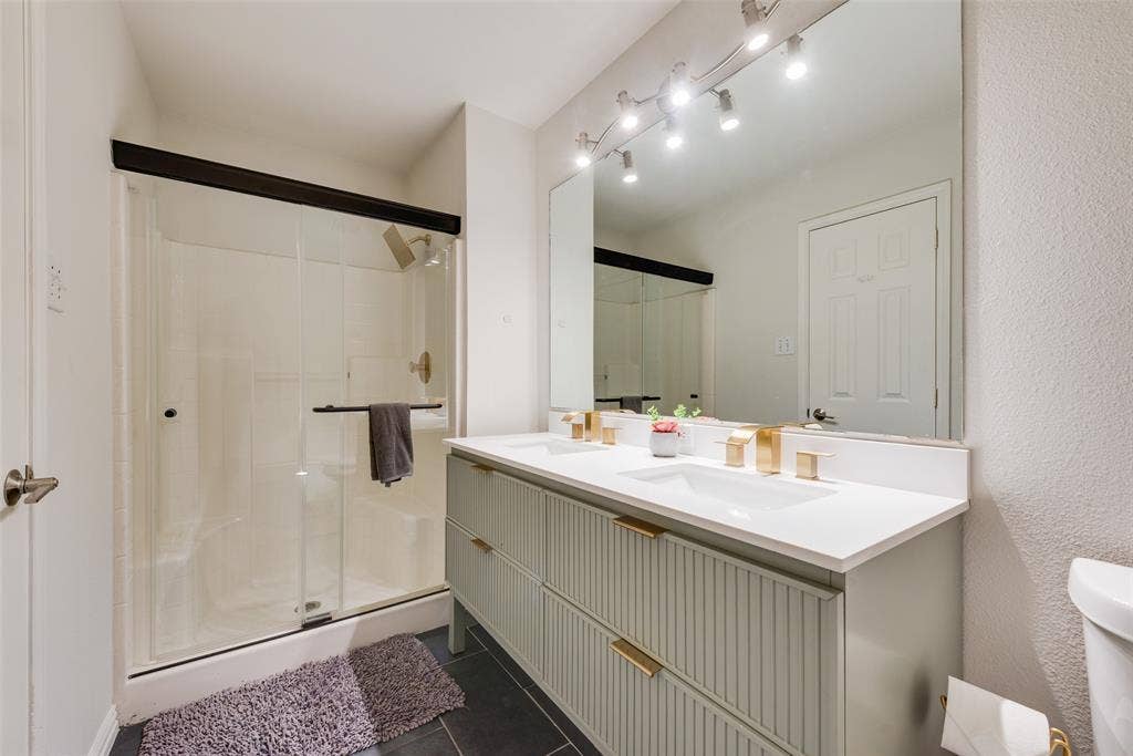 4104 Creekmeadow Drive, Carrollton, TX 75010 Double vanity featuring a fluted-style cabinet base, stone countertop, integrated rectangular sinks, and metallic finish hardware, complemented by a walk-in shower with a glass enclosure