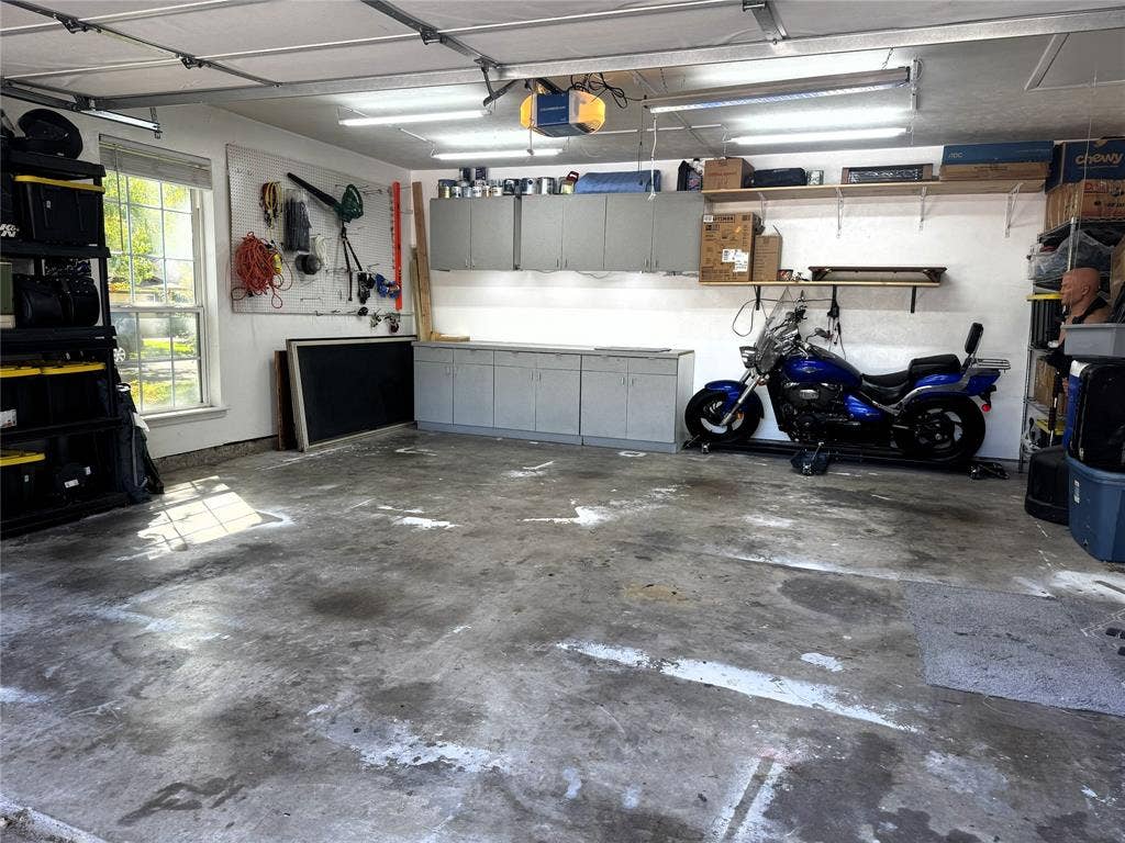 Two-car garage featuring wall-mounted upper and lower cabinetry, open shelving, fluorescent overhead lighting, and a single-pane window
