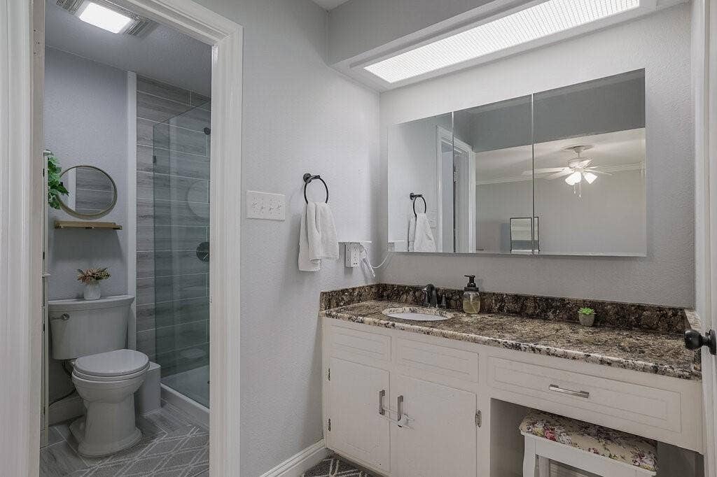 Bathroom featuring a vanity with stone-patterned countertop, white cabinetry with integrated makeup desk, tri-fold medicine cabinet, recessed lighting, and a glass-enclosed shower with tile surround