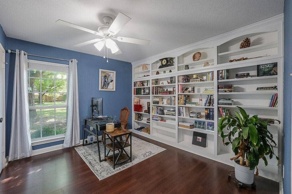 Wall-to-wall built-in shelving with arched detailing, wood-finish flooring, white ceiling fan with integrated lighting, and large window with natural light