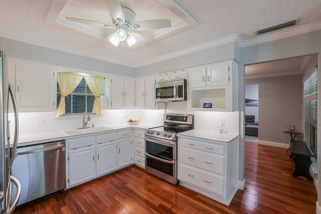 Kitchen featuring white cabinetry, wood-finish flooring, stainless steel appliances, a recessed ceiling tray with a central fan, and under-cabinet lighting