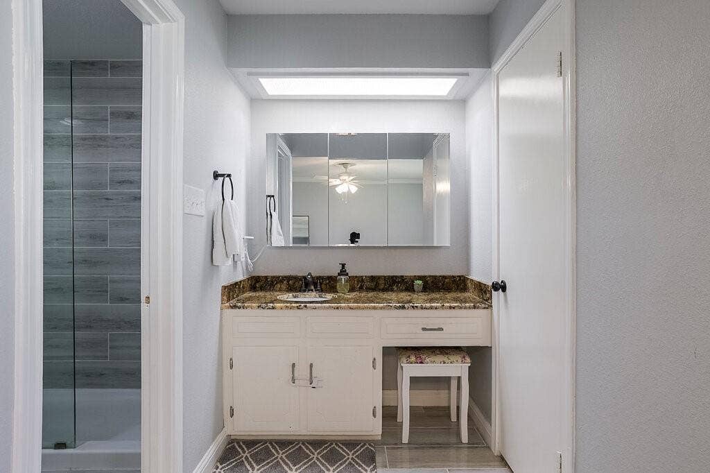 Built-in white vanity with stone-style countertop and integrated sink, recessed lighting, tri-panel medicine cabinet, and glass-enclosed shower with horizontal wall tile
