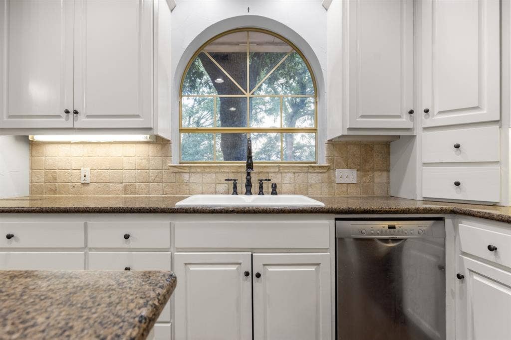 207 Westlane Circle, Woodway, TX 76712 Kitchen featuring white shaker-style cabinetry, stone-finish countertops, textured tile backsplash, arched window with decorative muntins, and a dual-basin white sink with a dark-finish faucet