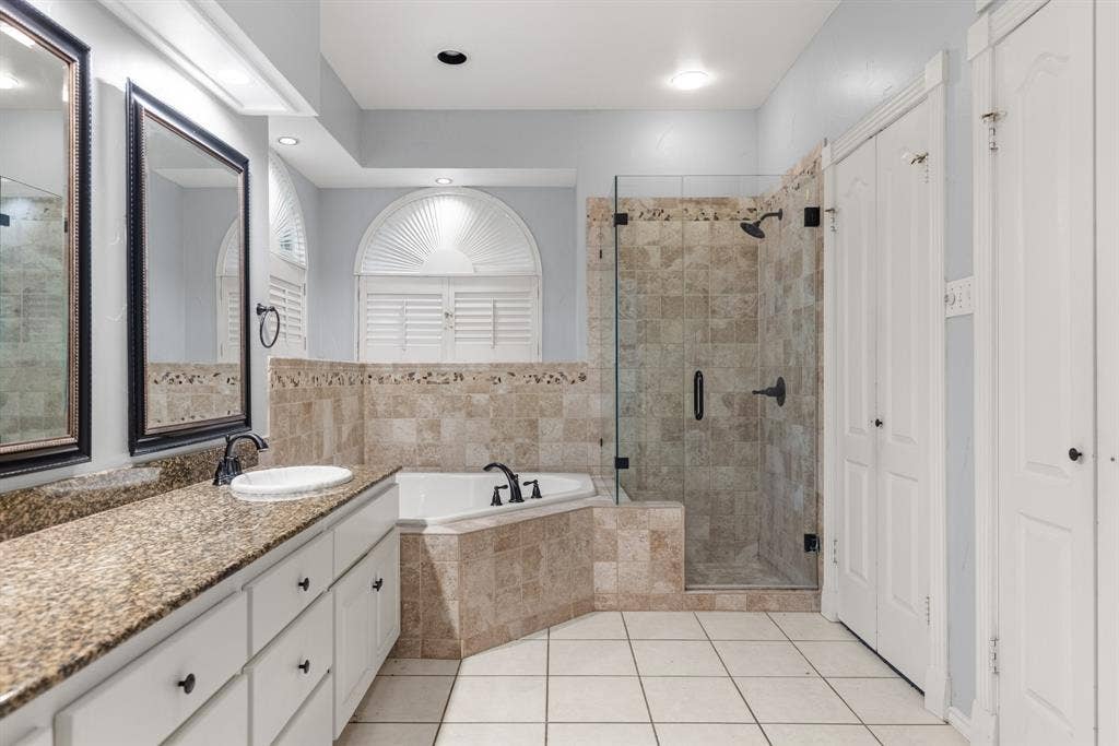 207 Westlane Circle, Woodway, TX 76712 Dual-sink vanity with stone countertop, corner soaking tub with tile surround, glass-enclosed walk-in shower, and arched window with plantation shutters