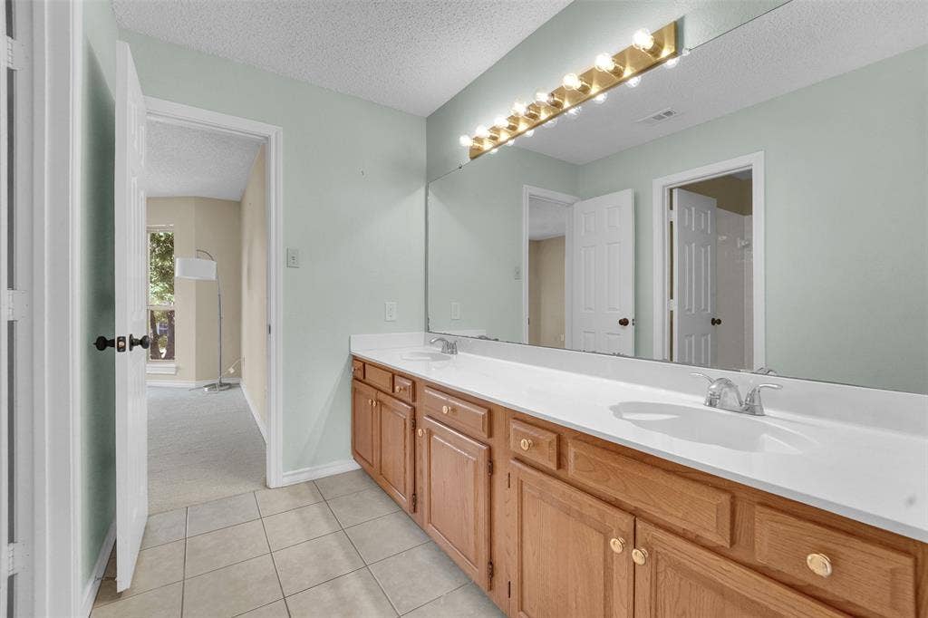1920 Keystone Drive, Plano, TX 75075 Dual-sink vanity with wood-finish cabinetry, solid-surface countertop, full-width mirror, and multi-bulb light fixture, complemented by tiled flooring