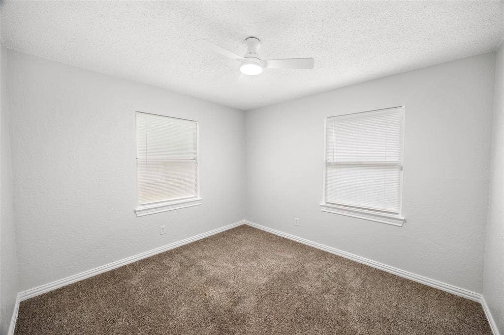 1605 Proctor Drive, Grand Prairie, TX 75051 Room featuring light grey walls, brown carpet, two windows with blinds, white trim, and a white ceiling fan