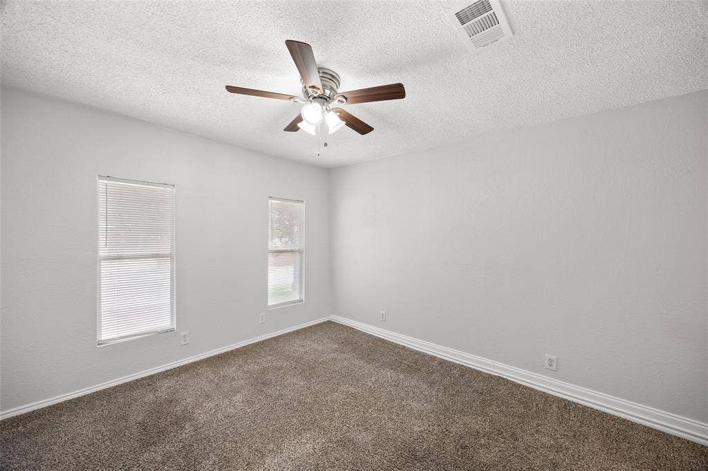 1605 Proctor Drive, Grand Prairie, TX 75051 Spacious room featuring neutral carpeting, light gray walls, a textured ceiling, two windows with blinds, and a ceiling fan with integrated lighting