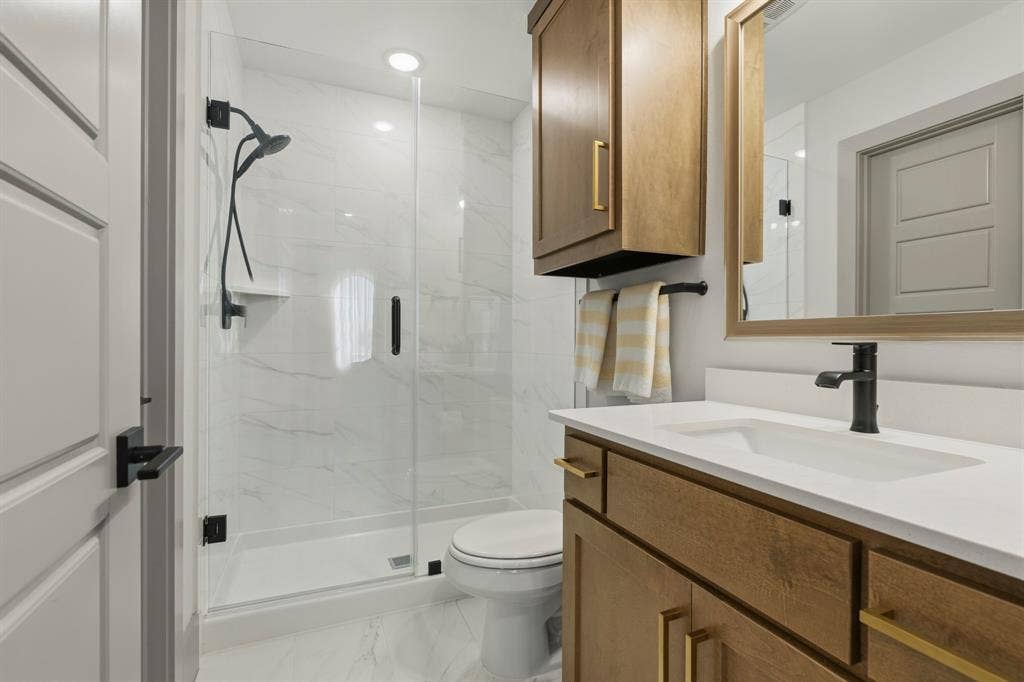 12173 Deer Trail, Frisco, TX 75035 Bathroom featuring a glass-enclosed shower with large-format wall tile, wood-finish vanity cabinetry with gold-toned hardware, quartz-composite countertop, and matte black plumbing fixtures