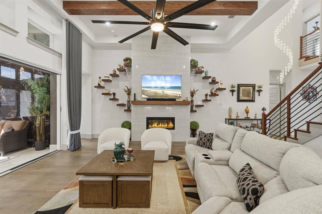 12173 Deer Trail, Frisco, TX 75035 High-ceilinged living area featuring a fireplace with a full-height tile surround, exposed wooden beam, large-scale multi-blade ceiling fan, and staircase with metal railing
