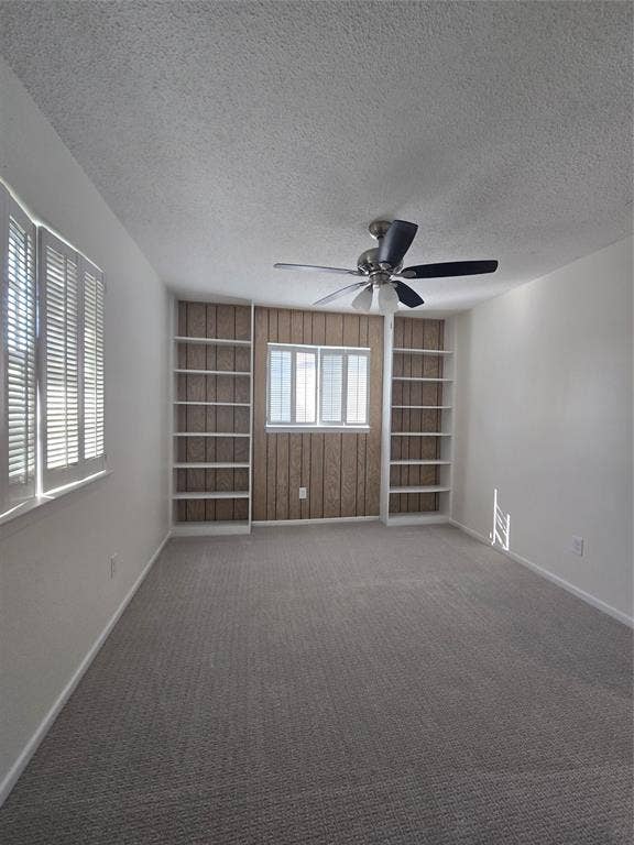 Room featuring neutral carpeting, built-in floor-to-ceiling shelving, wood-panel accent wall, dual windows with plantation shutters, and a ceiling fan with integrated lighting