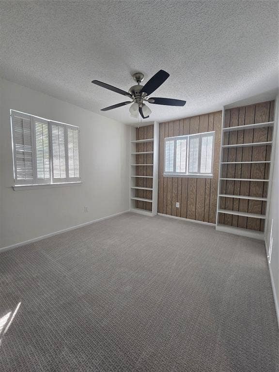 Room featuring wall-to-wall carpeting, ceiling fan, wood-paneled accent wall with built-in shelving, and white plantation-style shutters