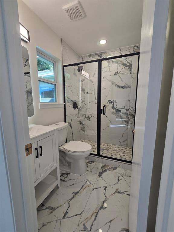 Bathroom featuring large-format patterned tile flooring, a walk-in shower with black-framed glass enclosure, stone-finish wall tile, pebble-style shower floor, and a white cabinet vanity