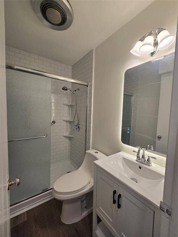 Bathroom featuring a white vanity with an integrated sink, a rectangular mirror with integrated perimeter lighting, a walk-in shower with white subway tile surround, and wood-finish flooring
