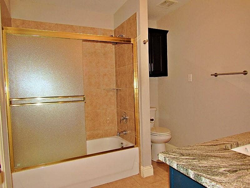 313 Timbercreek Court, Princeton, TX 75407 Bathroom featuring a tub-shower combination with floor-to-ceiling tile surround, gold-toned sliding glass enclosure, stone-surface countertop, and built-in dark wood-finish cabinetry