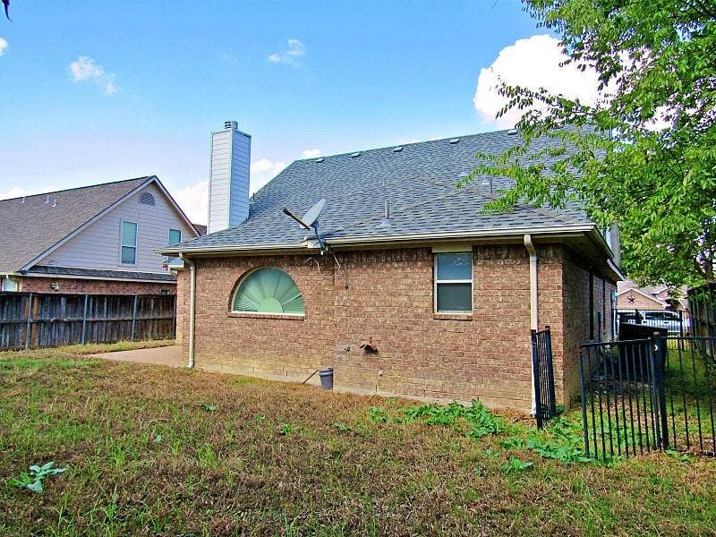 313 Timbercreek Court, Princeton, TX 75407 Brick exterior featuring a tall chimney stack, arched window, asphalt shingle roof, and metal perimeter fencing